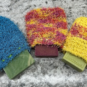 May include: Three bars of soap wrapped in colorful, textured, crocheted fabric. The soap on the left is green, the middle soap is purple, and the soap on the right is green with the word "AUSTRALIA" printed on it.