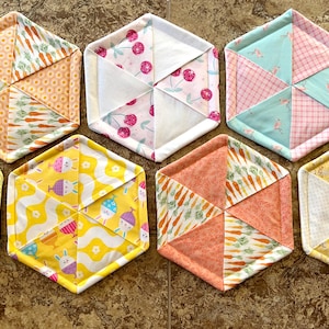 May include: Six hexagonal fabric coasters with different patterns. The coasters are made of patchwork fabric with various colors and designs, including carrots, cherries, bunnies, and geometric patterns.