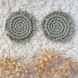 May include: Two round, light green crocheted coasters with a fringe border. The coasters are on a white fluffy surface.