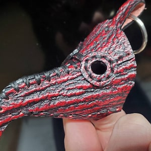 May include: A red and black leather plague doctor mask keychain. The mask has a long, pointed beak and a small eye hole. The keychain is attached to a silver ring.