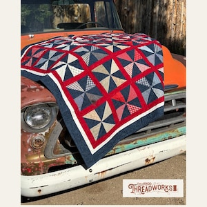 Op de afbeelding: Een kleurrijk quilt met een windmolenontwerp in rood, wit en blauw, gedrapeerd over de motorkap van een oranje vintage truck. De quilt heeft een marineblauwe rand met een witte streep. De tekst "Liberty Pinwheel Quilt Pattern" is inbegrepen.