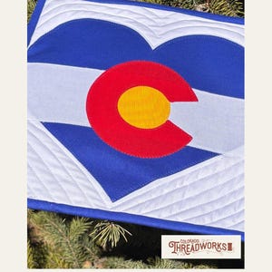 May include: A quilted textile featuring the Colorado state flag design. The design includes a red 'C' with a yellow center on a blue background, a white stripe, and white heart shapes. The text "COLORADO HEARTSTRINGS QUILT PATTERN" is at the bottom.