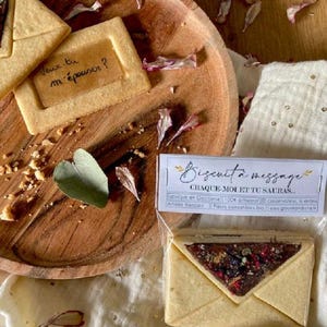 Pode incluir: Um close-up de um biscoito em forma de envelope com uma mensagem dentro. O biscoito está sobre uma superfície de madeira com um pequeno saco de biscoitos no fundo. O saco tem o texto "Biscuit à message CRAQUE MOI ET TU SAURAS.....". O biscoito é uma forma divertida e original de compartilhar uma mensagem com alguém especial.