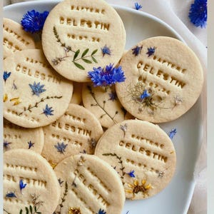 Peut inclure: Gros plan sur plusieurs biscuits ronds de couleur brun clair décorés de fleurs pressées et du texte "Bapteme Celestin 08.07.24".