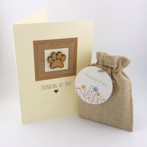 Op de afbeelding: Een condoleancekaart met een houten pootprint op een jute achtergrond. De kaart zegt "Thinking of you" met een klein hartje. Een klein jute zakje met een label dat "Thinking of you" zegt met een bloemenpatroon.
