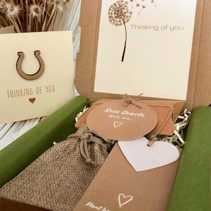 May include: A brown cardboard gift box containing a burlap drawstring pouch, a round tag labeled Rose Quartz With love, a heart-shaped tag, and a card with a dandelion illustration and the text Thinking of you, plus a separate matching card.