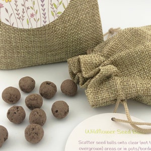 Op de afbeelding: Een klein jute zakje gevuld met bruine wilde bloemen zaadballen. Het zakje heeft een trekkoord sluiting en is vastgebonden met een wit lint. Het zakje ligt op een wit oppervlak. Een wit kaartje met de tekst "Wildflower Seed Balls" ligt naast het zakje. Het kaartje bevat instructies over hoe de zaadballen te planten.