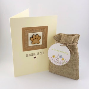 Op de afbeelding: Een condoleancekaart met een houten pootprint op een jute achtergrond. De kaart zegt "Thinking of You" met een hart. Een kleine jute zak met een label dat "In loving memory" zegt met bloemen.