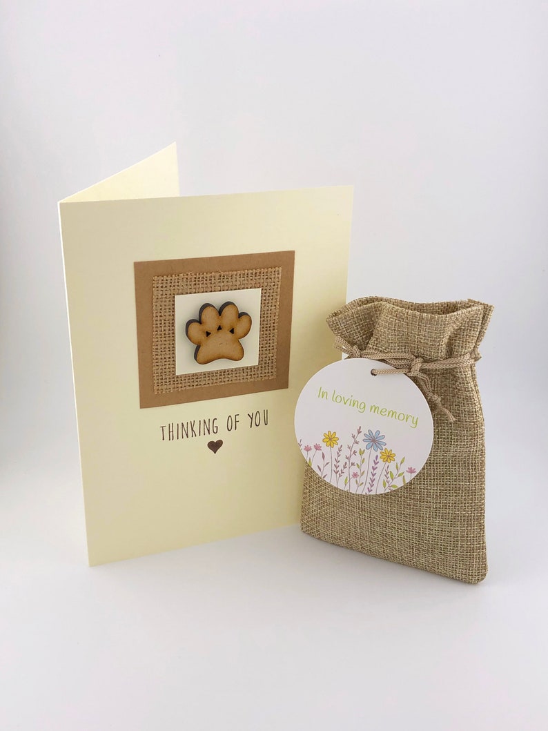 Op de afbeelding: Een condoleancekaart met een houten pootprint op een jute achtergrond. De kaart zegt "Thinking of You" met een hart. Een kleine jute zak met een label dat "In loving memory" zegt met bloemen.