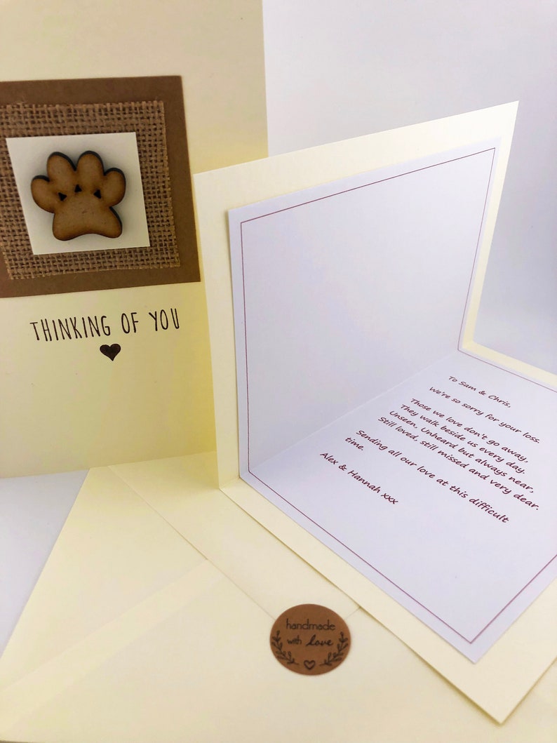 Op de afbeelding: Een condoleancekaart met een houten pootprint op een jute achtergrond. De kaart heeft de tekst "Thinking of You" met een hart. De binnenkant van de kaart heeft een handgeschreven condoleancebericht.