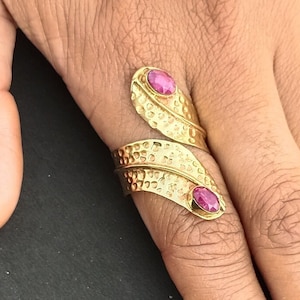 May include: A gold-colored ring with a leaf design, featuring two oval-shaped, pink gemstones. The ring has a textured surface and wraps around the finger. The gemstones are set in a bezel setting.