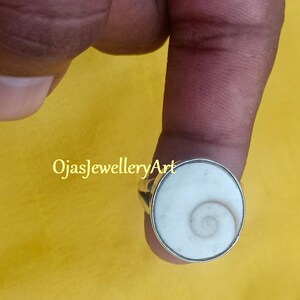 Puede incluir: Un anillo de plata con una incrustación de concha blanca que presenta un patrón en espiral. El anillo está engastado en una banda de plata y se lleva en un dedo.