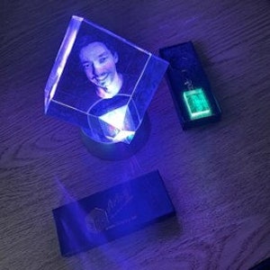 Puede incluir: Un cubo de cristal transparente y brillante con un retrato en su interior, iluminado por una luz azul. Junto a &eacute;l, una caja rectangular y un llavero con un brillo verde. Sobre una superficie de madera.
