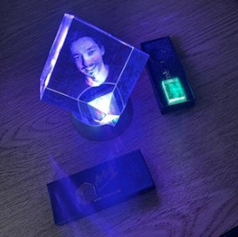Puede incluir: Un cubo de cristal transparente y brillante con un retrato en su interior, iluminado por una luz azul. Junto a &eacute;l, una caja rectangular y un llavero con un brillo verde. Sobre una superficie de madera.