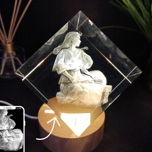 Puede incluir: Una escultura de cristal transparente en forma de diamante con una imagen grabada de una mujer sentada. La escultura est&aacute; montada sobre una base de madera con una luz c&aacute;lida y brillante. La imagen es de una mujer con un vestido fluido, sosteniendo un instrumento musical.