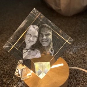 Puede incluir: Un bloque de cristal transparente en forma de diamante con una imagen grabada con l&aacute;ser de dos personas sonriendo. El cristal se asienta sobre una base de madera con una luz c&aacute;lida que ilumina la imagen grabada. Un cable de alimentaci&oacute;n se extiende desde la base.
