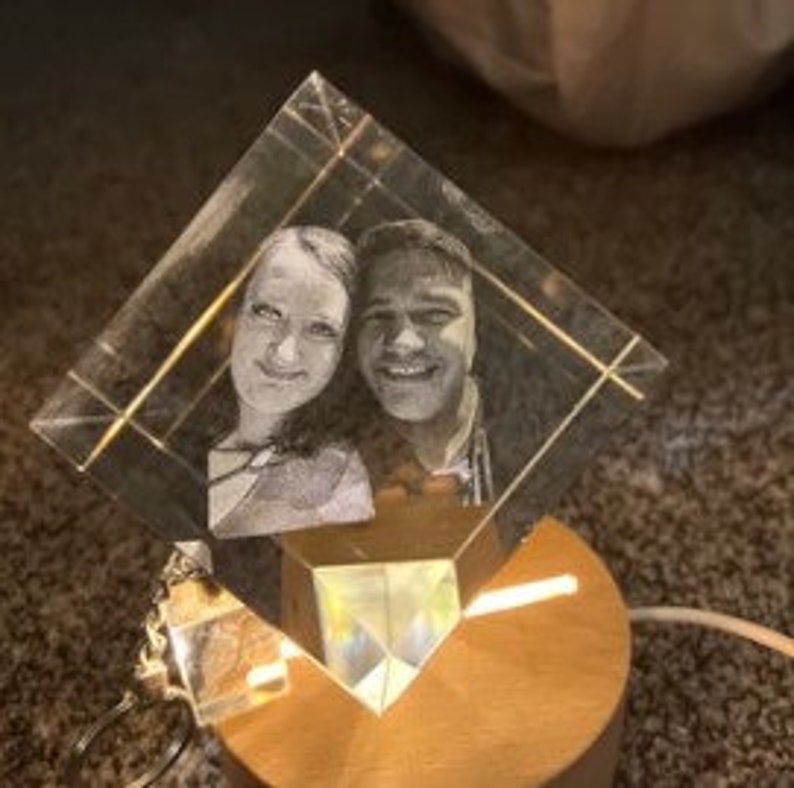 Puede incluir: Un bloque de cristal transparente en forma de diamante con una imagen grabada con l&aacute;ser de dos personas sonriendo. El cristal se asienta sobre una base de madera con una luz c&aacute;lida que ilumina la imagen grabada. Un cable de alimentaci&oacute;n se extiende desde la base.