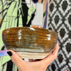 May include: A handmade ceramic bowl with a textured surface. The bowl features a blend of brown, orange, and blue hues, with a lighter beige base. The interior is smooth and glossy, while the exterior has a rough, rustic appearance.