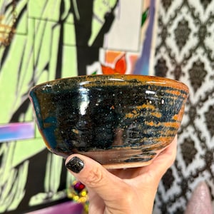 May include: A handmade ceramic bowl with a dark blue and orange glaze. The bowl has a round shape and a speckled texture. The rim is a lighter orange color. The bowl is held in a hand.
