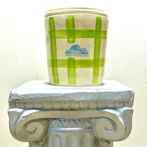 Puede incluir: Una taza de cerámica blanca con un patrón de cuadros verde y blanco y diseños de nubes azules. La taza está sentada sobre un pedestal plateado.