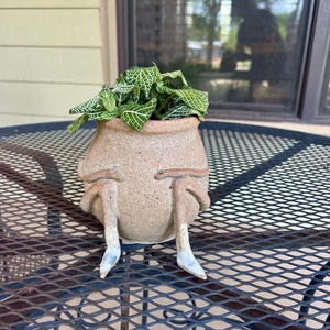 May include: A ceramic planter shaped like a person with legs and feet, holding a green plant with white veins. The planter is sitting on a black metal table.