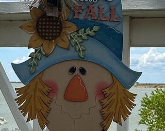 Può includere: Decorazione di spaventapasseri in legno con cappello blu, girasole e la scritta "Hello Fall". Lo spaventapasseri ha una sciarpa rossa e capelli di paglia. Un fiocco decorativo è in cima. Lo sfondo mostra uno specchio d'acqua e un cielo blu.