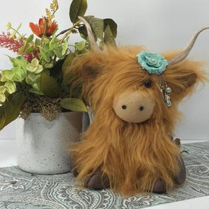 May include: A plush brown Highland cow toy with a blue flower and beaded jewelry on its head. The toy is sitting on a patterned surface.