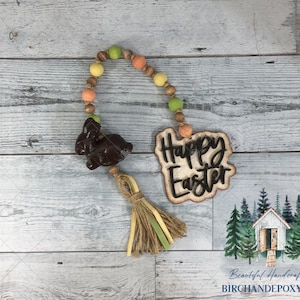 May include: A wooden Easter garland with a brown bunny charm, a wooden sign that says "Happy Easter", and a brown, yellow, and green tassel. The garland is made of wooden beads in shades of brown, yellow, green, and orange.