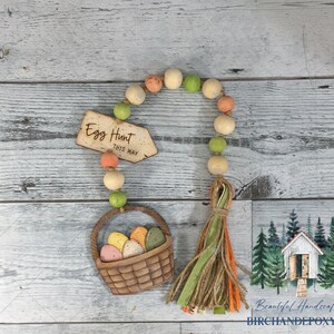 May include: A wooden Easter egg hunt sign with the text "Egg Hunt This Way" hanging from a beaded garland with a basket of colorful eggs and a green, brown, and orange tassel.