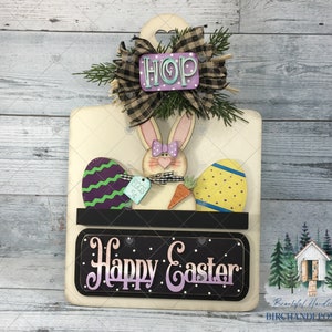 May include: A white wooden sign with a bunny rabbit, eggs, and a carrot. The sign says "Happy Easter" and has a black shelf with a sign that says "Eggs 5¢". There is a purple and green bow with the word "HOP" on it.