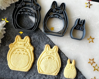 May include: A set of six cookie cutters in the shape of Totoro characters. The cutters are made of black metal and are arranged on a gray surface. There are also several cookies baked in the shape of Totoro characters.