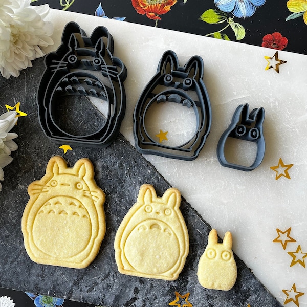 May include: A set of six cookie cutters in the shape of Totoro characters. The cutters are made of black metal and are arranged on a gray surface. There are also several cookies baked in the shape of Totoro characters.
