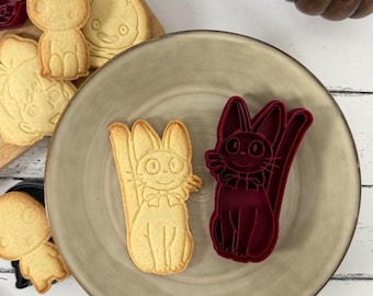 May include: Two cookie cutters in the shape of cats. One is red and the other is a light brown. The red cookie cutter is in the shape of a cat with a bow tie. The light brown cookie cutter is in the shape of a cat with a bow tie and a tail.