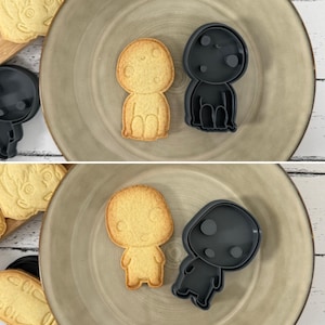 Peut inclure: Deux emportes-pièces et deux biscuits en forme de petit personnage de dessin animé rond avec un visage souriant. Les emportes-pièces sont noirs et les biscuits sont brun clair.