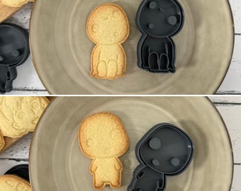 May include: Two cookie cutters and two cookies in the shape of a small, round, cartoon character with a smiling face. The cookie cutters are black and the cookies are light brown.