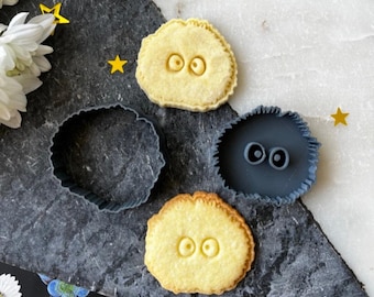 May include: Three cookie cutters in the shape of a round creature with two eyes. The cutters are made of dark gray plastic. There are three cookies next to the cutters, each with a round creature design and two eyes.