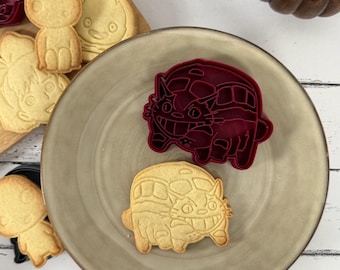 May include: Two cookie cutters in the shape of a cat bus from the movie My Neighbor Totoro. The cutters are red and beige. The cutters are on a beige plate.