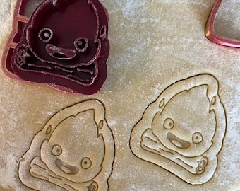 May include: Two cookie cutters, one red and one brown, shaped like a cartoon character with a large head and a small body. The character has large eyes, a small mouth, and a large head of hair.
