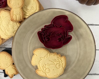 May include: Two red plastic cookie cutters shaped like cartoon characters with large eyes and hair. The cutters are on a brown plate with a cookie cut out using one of the cutters.