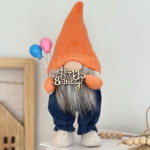 Puede incluir: Una muñeca de gnomo caprichosa con un gorro de punto naranja y pantalones vaqueros azules. El gnomo sostiene un letrero de madera que dice "Happy Birthday" y dos globos. Tiene una larga barba gris y zapatos blancos. Una decoración festiva de cumpleaños.