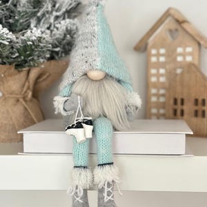 May include: A whimsical gnome doll with a gray and light blue knit hat and beard. The gnome is wearing light blue knit legs and gray knit boots with white faux fur trim. The gnome is holding a pair of miniature ice skates.