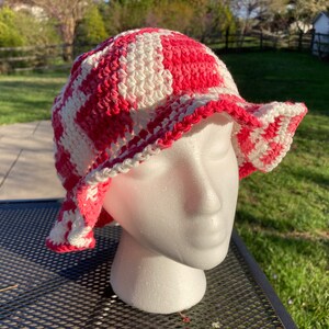 May include: A crocheted bucket hat with a pink and white checked pattern. The hat is made with a soft, textured yarn and has a wide brim.