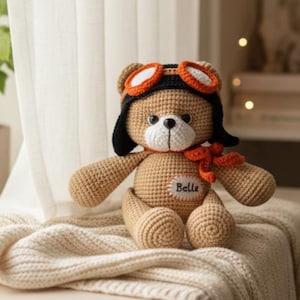 Könnte beinhalten: Ein gehäkelter beigefarbener Teddybär mit einer schwarz-orangen Fliegermütze samt Schutzbrille und einem passenden orangefarbenen Schal. Der Bär hat eine weiße Schnauze, schwarze Augen und einen weißen ovalen Aufnäher mit dem Namen Belle in Schwarz.