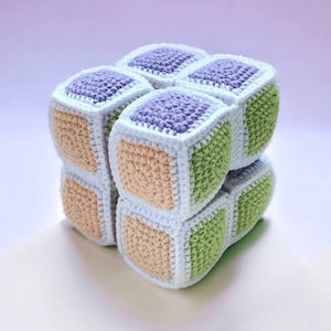 Può includere: Un giocattolo a forma di cubo all'uncinetto con una varietà di colori, tra cui viola, verde e beige. Il cubo è composto da quadrati più piccoli che sono cuciti insieme.