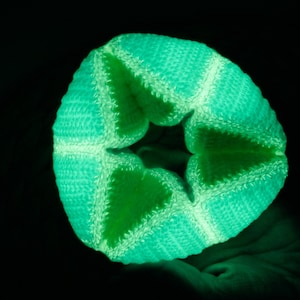 Könnte beinhalten: Ein grünes, gehäkeltes sternförmiges Objekt leuchtet im Dunkeln. Das Objekt besteht aus einem weichen, strukturierten Material und hat eine dreidimensionale Form.