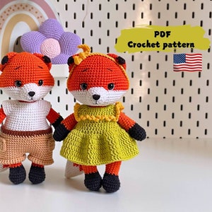Op de afbeelding: Twee gehaakte vossenpoppen, één in een wit shirt en bruine shorts, de andere in een gele jurk. Beide hebben oranje vacht, zwarte accenten en witte gezichten. De afbeelding bevat de tekst "PDF Crochet pattern" en een kleine Amerikaanse vlag.