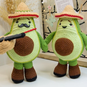 May include: Two crocheted avocado plushies wearing sombreros. One avocado is playing a small guitar and the other is smiling.