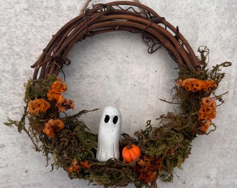Puede incluir: Una corona circular hecha de ramas marrones entrelazadas, decorada con musgo verde y pequeñas flores naranjas. Una figura de fantasma blanco con dos ojos negros y una pequeña calabaza naranja están situadas en la parte inferior central.