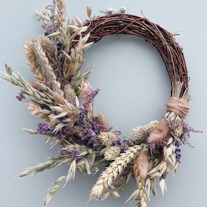 Op de afbeelding: Een gedroogde bloemenkrans met een bruine takkenbasis. De krans is versierd met gedroogde lavendel, haver en andere gedroogde grassen.