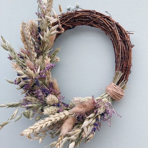 Op de afbeelding: Een krans van gedroogde bloemen met een bruine wijnstokbasis. De krans is versierd met gedroogde lavendel, tarwe en andere grassen.